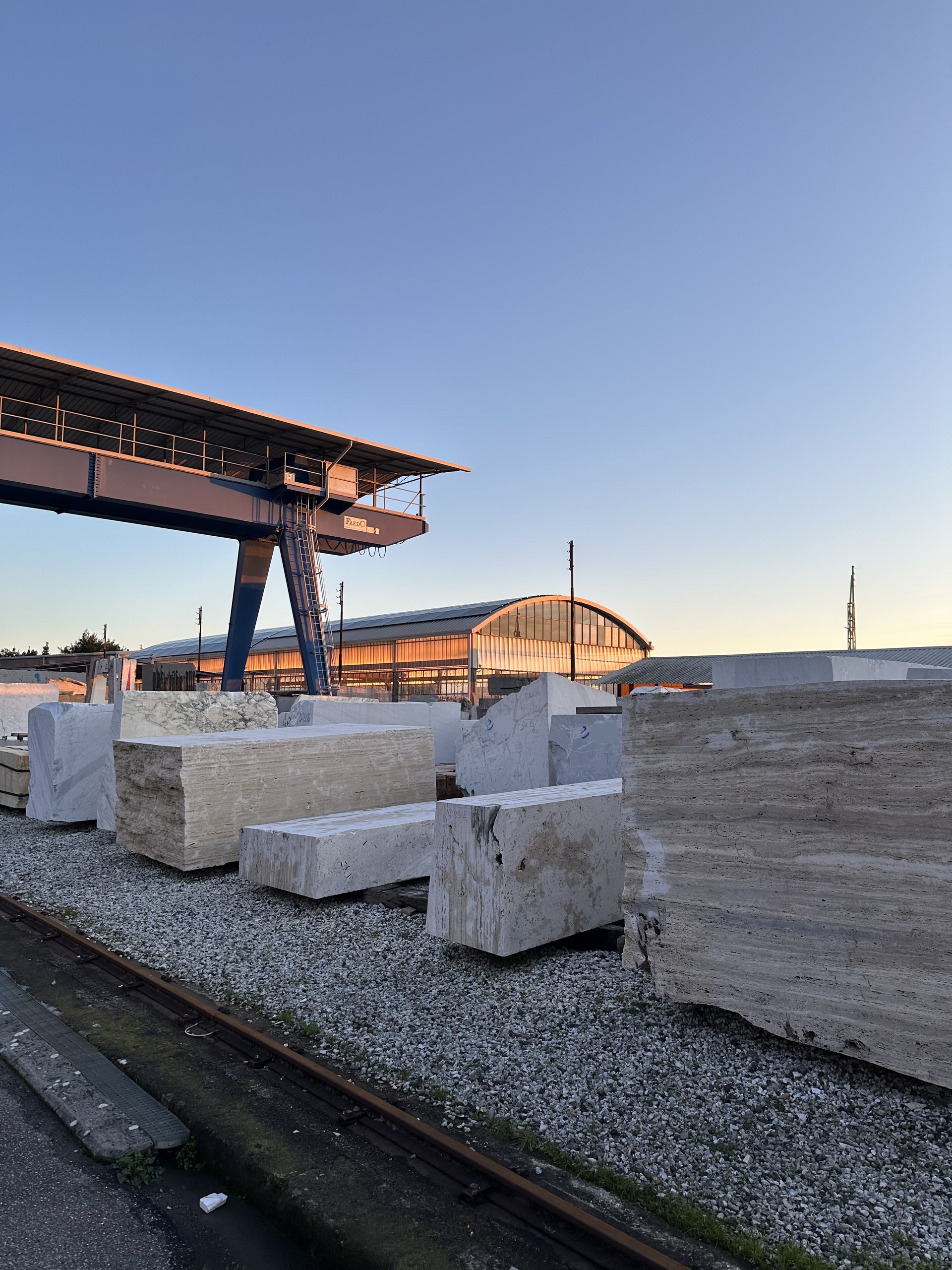 Quarry yard at golden hour — rows of raw travertine blocks and stone slabs beneath a large gantry crane, with a warehouse shed and railway tracks behind. Novo Marble stone, from the quarry to the project.